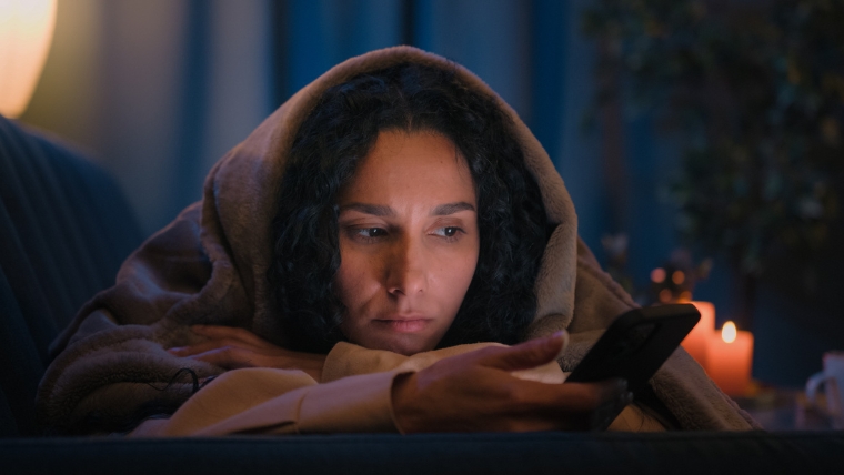 Woman of color on couch under blanket scrolling phone with a sad expression | The Psychology of Comparison Why We Do It and How to Stop | Mindful Health Solutions