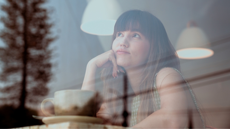 Young woman at a coffee shop looking thoughtfully out a window | Unlock the Power of Reflection and Set Powerful Mental Health Goals | Mindful Health Solutions