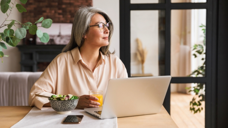 Mature woman sitting at a table with a salad and computer, looking off in the distance thinking | TMS vs. Traditional Pain Management Which Is Right for Your Chronic Pain | Mindful Health Solutions